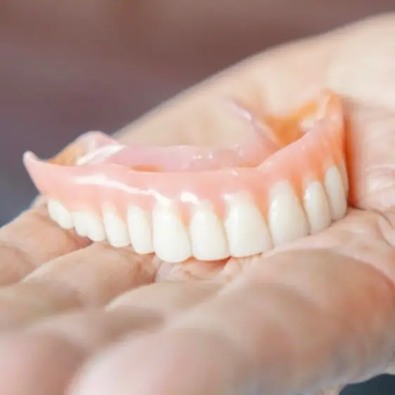 Hand holding realistic custom-fit dentures beside text highlighting patient benefits: eat, smile, and speak with confidence again in D. Dentist Dental Clinic.